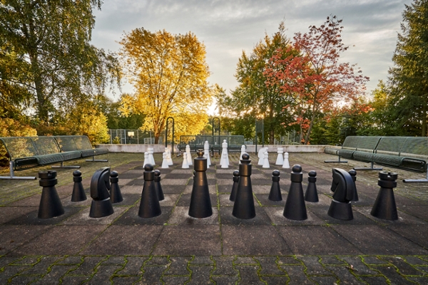 Gartenschach mit den schwarzen Figuren im Vordergrund umgeben von Sitzbänken und Bäumen
